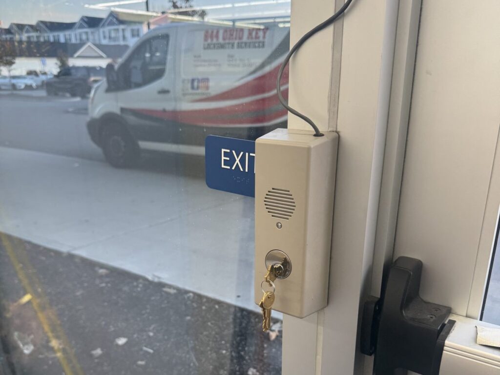 New mortise lock in a Detex exit alarm with its key, a spare key and the alarm box key, beside an exit door in Grove City
